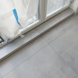 Szare płytki podłogowe, świeżo położone, z zabezpieczonym folią i taśmą oknem balkonowym, widoczne fugi.