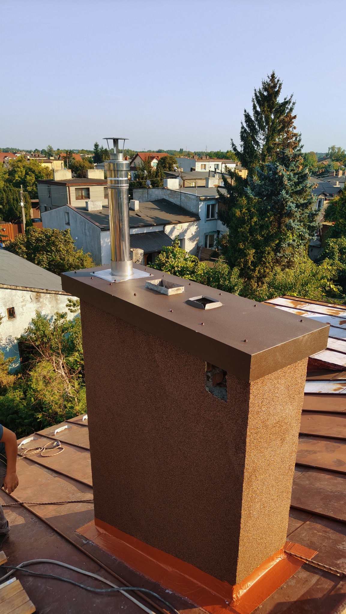 Otynkowany komin z metalowym daszkiem i stalowym wkładem kominowym, uszczelniony na dachu z blachy trapezowej, widok z góry na tle zabudowy miejskiej i zieleni.