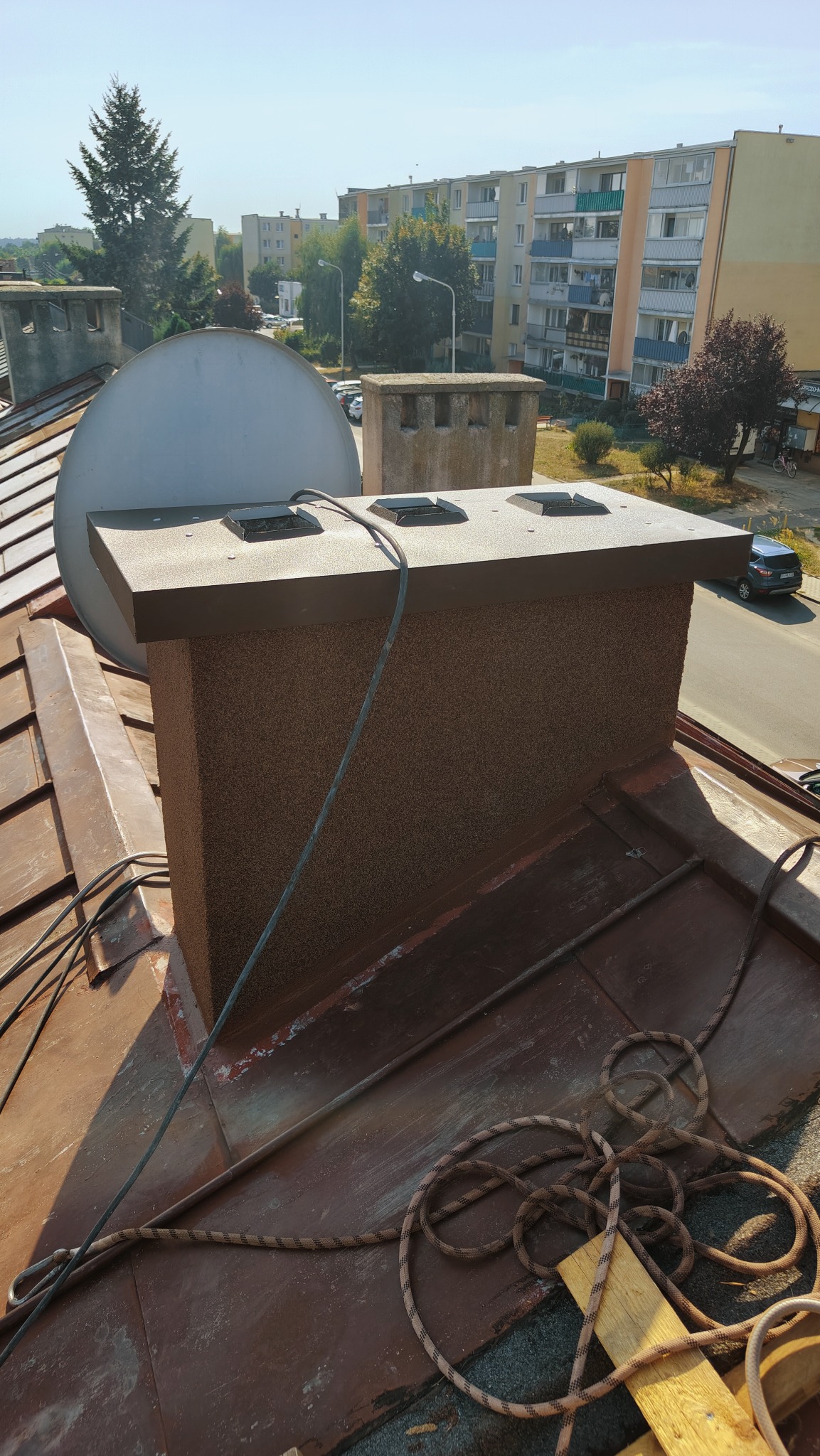 Widok na nowy komin z brązowym tynkiem strukturalnym na dachu z blachy, w tle zabudowa miejska i antena satelitarna.