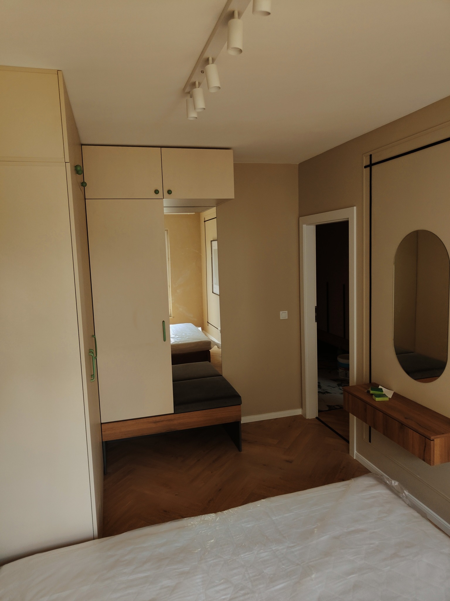 Ukończony remont pokoju w odcieniach beżu, widoczna szafa z siedziskiem, parkiet w jodełkę, lustro owalne oraz fragment zabezpieczonego folią łóżka.