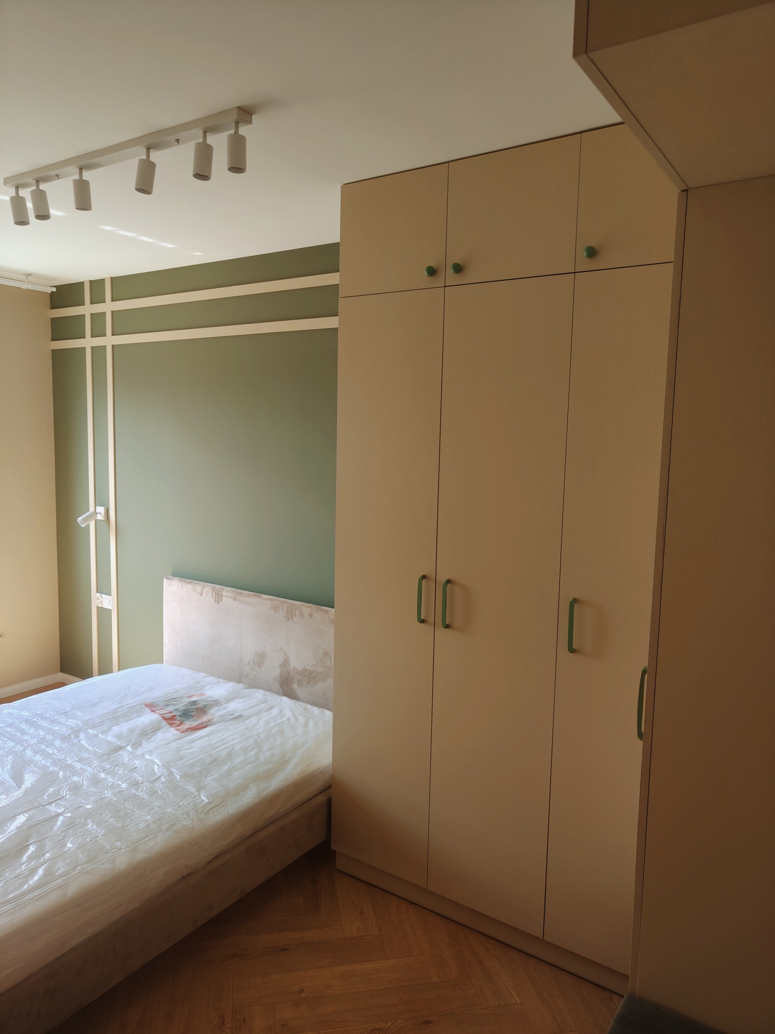 Pokój po odświeżeniu: ściana w odcieniach zieleni z dekoracyjnymi listwami, beżowa szafa z zielonymi uchwytami, łóżko z materacem w folii ochronnej, parkiet w jodełkę.
