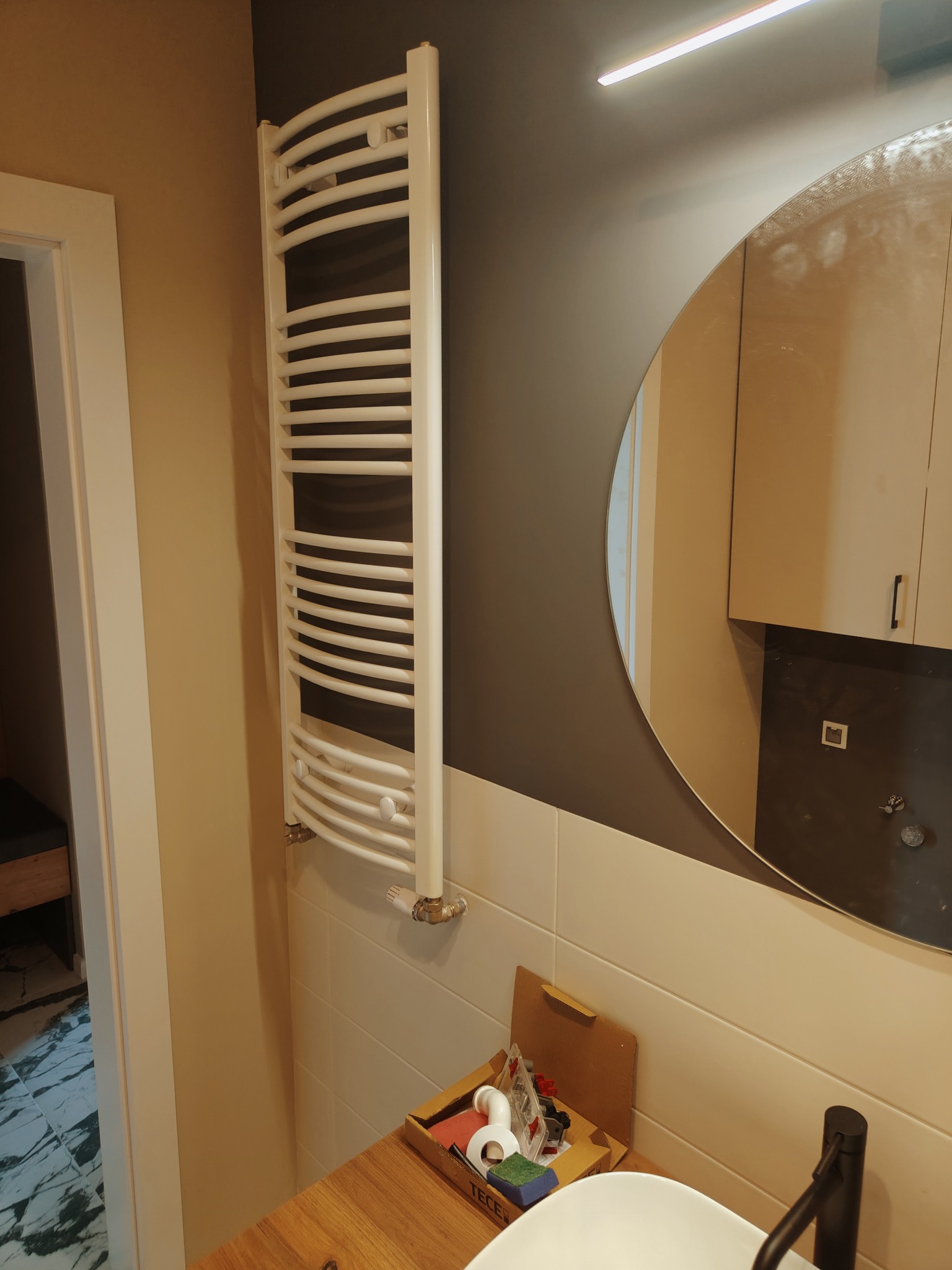 Nowoczesna łazienka w Łodzi po remoncie: biały grzejnik drabinkowy na ścianie z jasnych płytek, okrągłe lustro z widocznym odbiciem szafki, umywalka z czarną baterią i akcesoria łazienkowe...