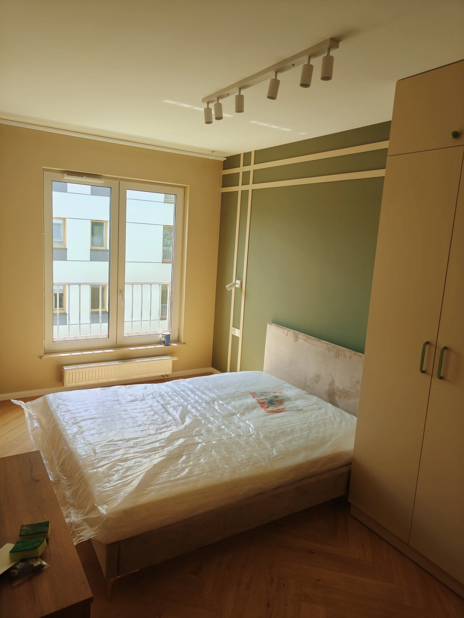 Pokój po remoncie w Łodzi: łóżko zabezpieczone folią, ściana w kolorze oliwkowym z dekoracyjnymi listwami, drewniana podłoga, duża szafa i widok z okna na blok.