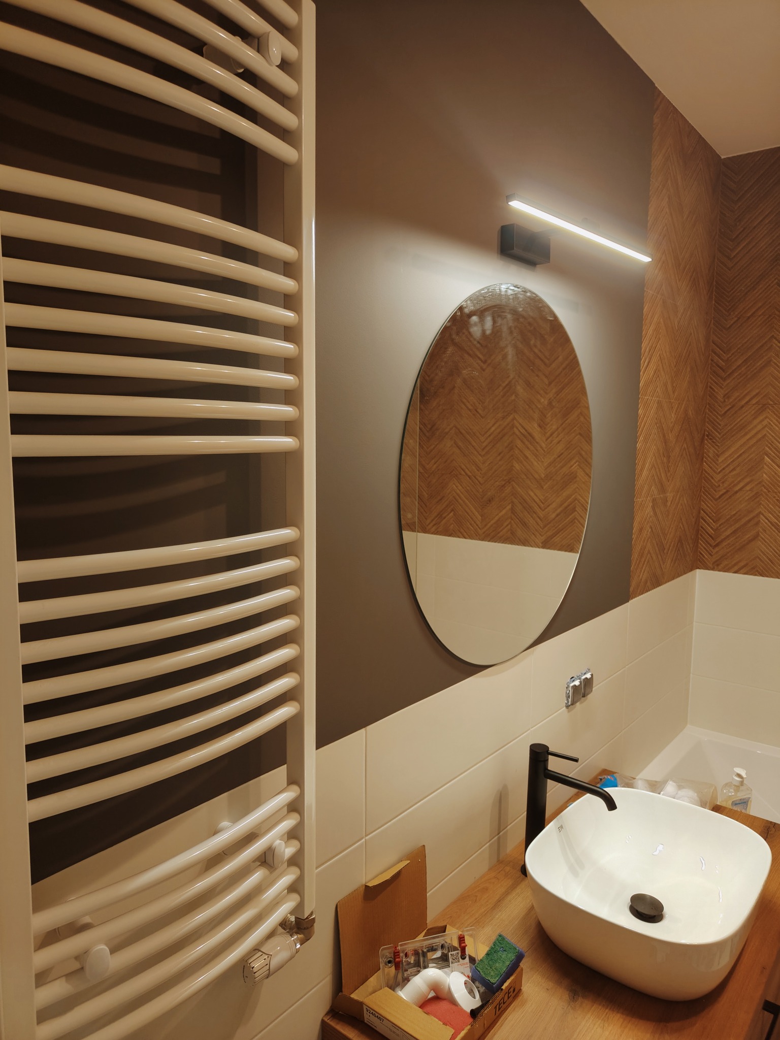 Nowoczesna łazienka po remoncie z owalnym lustrem, grzejnikiem drabinkowym i umywalką nablatową z czarną armaturą. Na blacie widoczne akcesoria montażowe i gąbki.