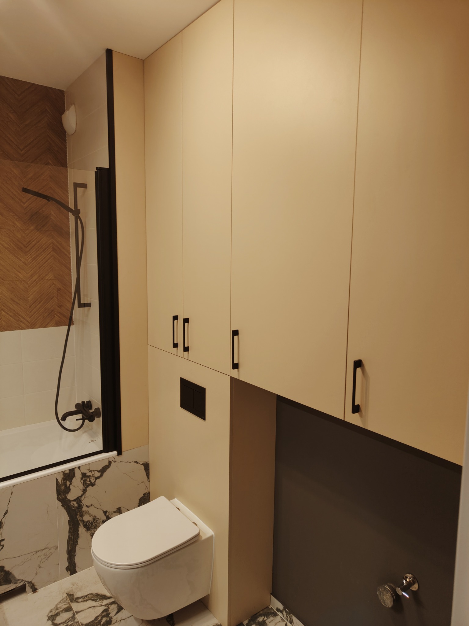Nowoczesna łazienka po remoncie: wisząca toaleta z podtynkową spłuczką, zabudowa meblowa z beżowymi frontami i czarnymi uchwytami, kabina prysznicowa z czarną ramą, ściany wykończone płytkami...