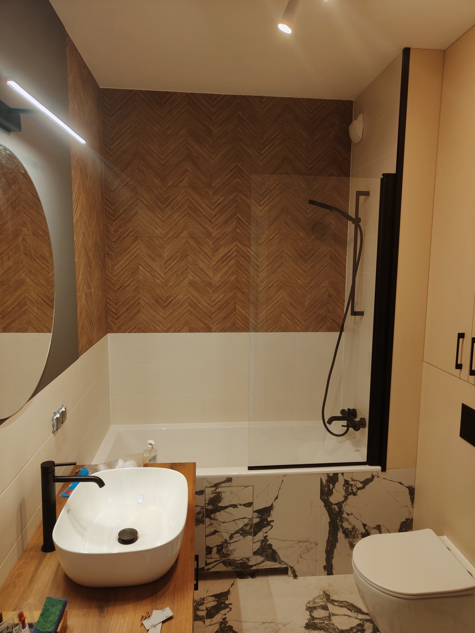 Nowoczesna łazienka z drewnopodobną mozaiką na ścianie, białą umywalką nablatową z czarną armaturą, oraz kabiną prysznicową z czarnymi profilami.