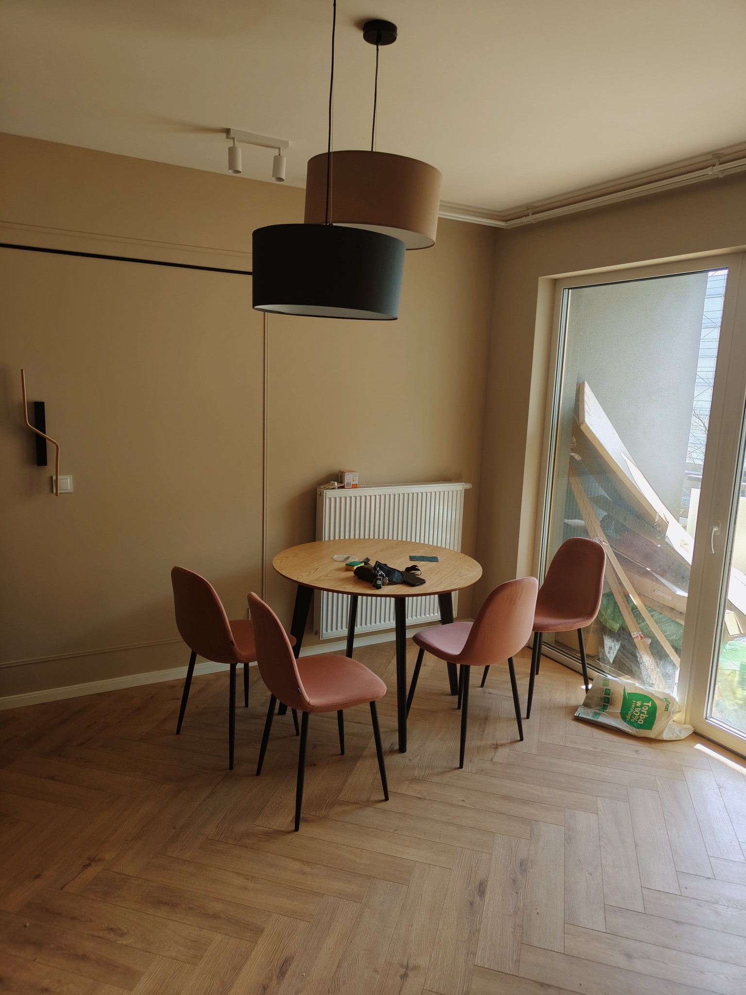 Jasne wnętrze z drewnianą podłogą ułożoną w jodełkę, okrągłym stołem otoczonym czterema różowymi krzesłami, wiszącymi lampami i oknem z widocznymi materiałami budowlanymi, sugerującymi prace...