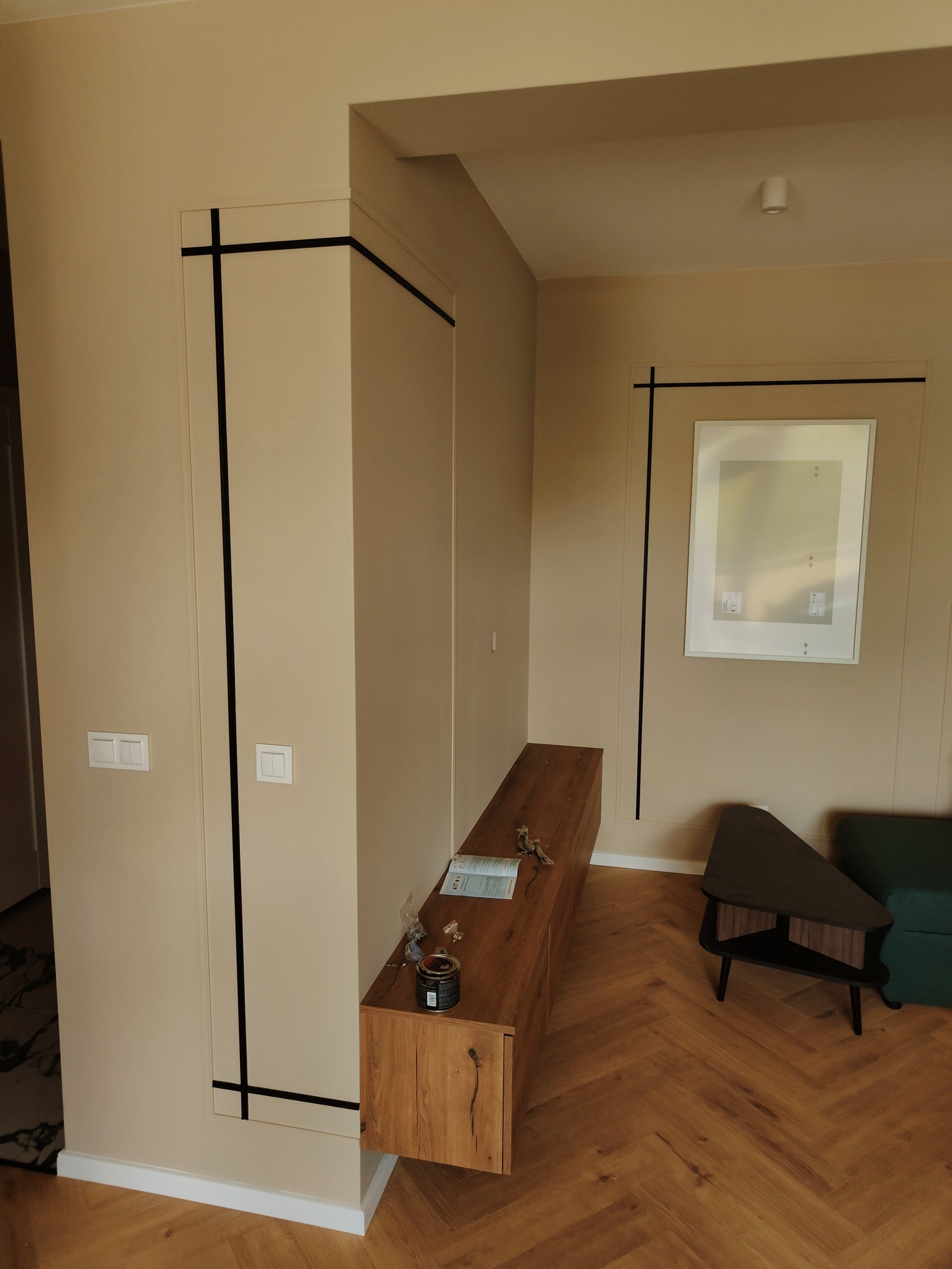 Beżowe wnętrze mieszkania po remoncie w Łodzi, z drewnianą podłogą w jodełkę, zabudową meblową z czarnymi akcentami oraz wiszącą szafką RTV z akcesoriami na blacie.