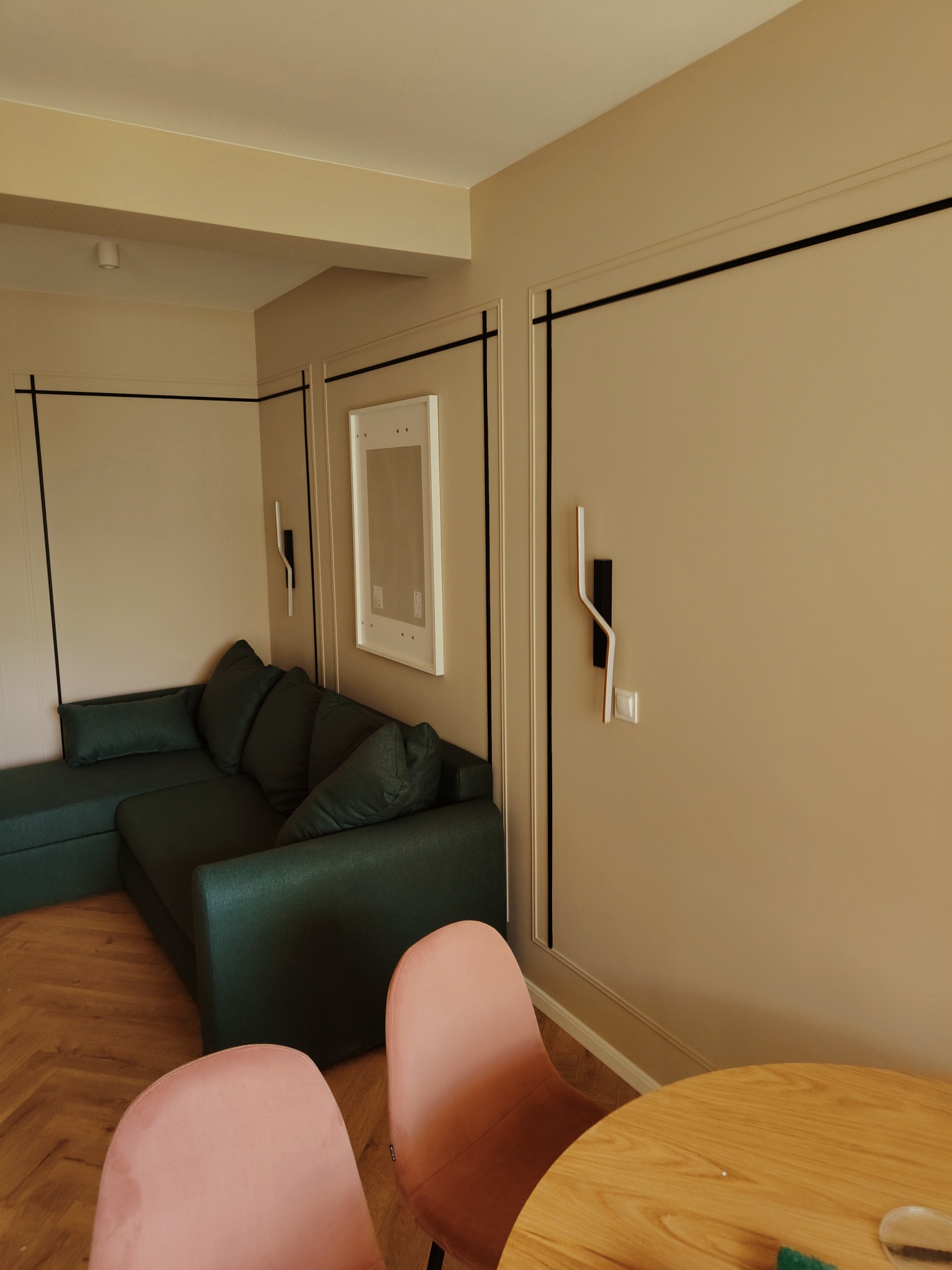 Beżowe ściany z czarnymi listwami dekoracyjnymi, narożna sofa w odcieniach zieleni, różowe krzesła i drewniany stół w wykończonym salonie po remoncie przy ul. Koczeńcowej w Łodzi.