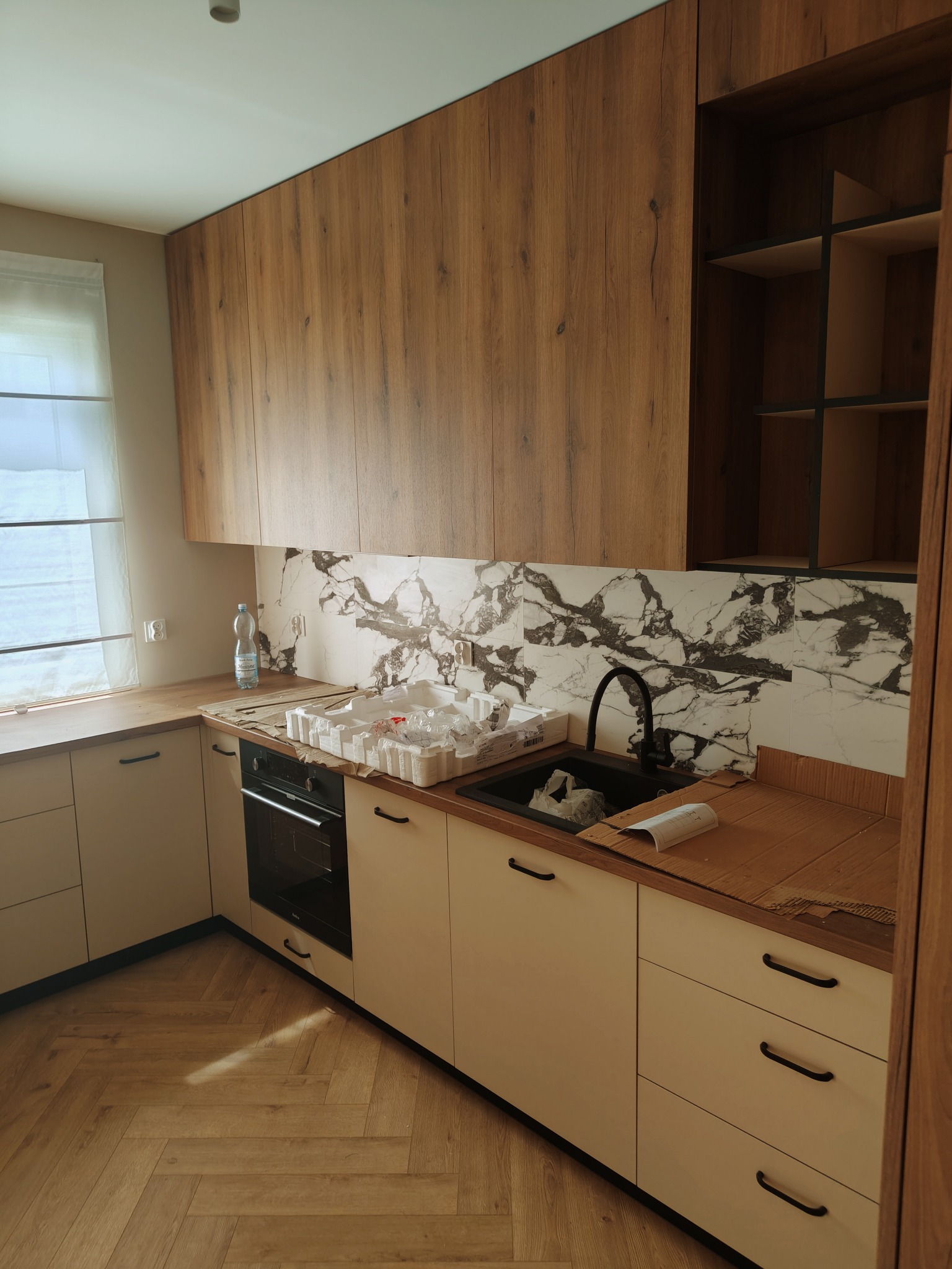 Nowoczesna kuchnia z drewnianymi szafkami górnymi i białymi dolnymi, marmurowym backsplash, czarnym zlewem i armaturą, oraz parkietem ułożonym w jodełkę; widoczne ślady po montażu.