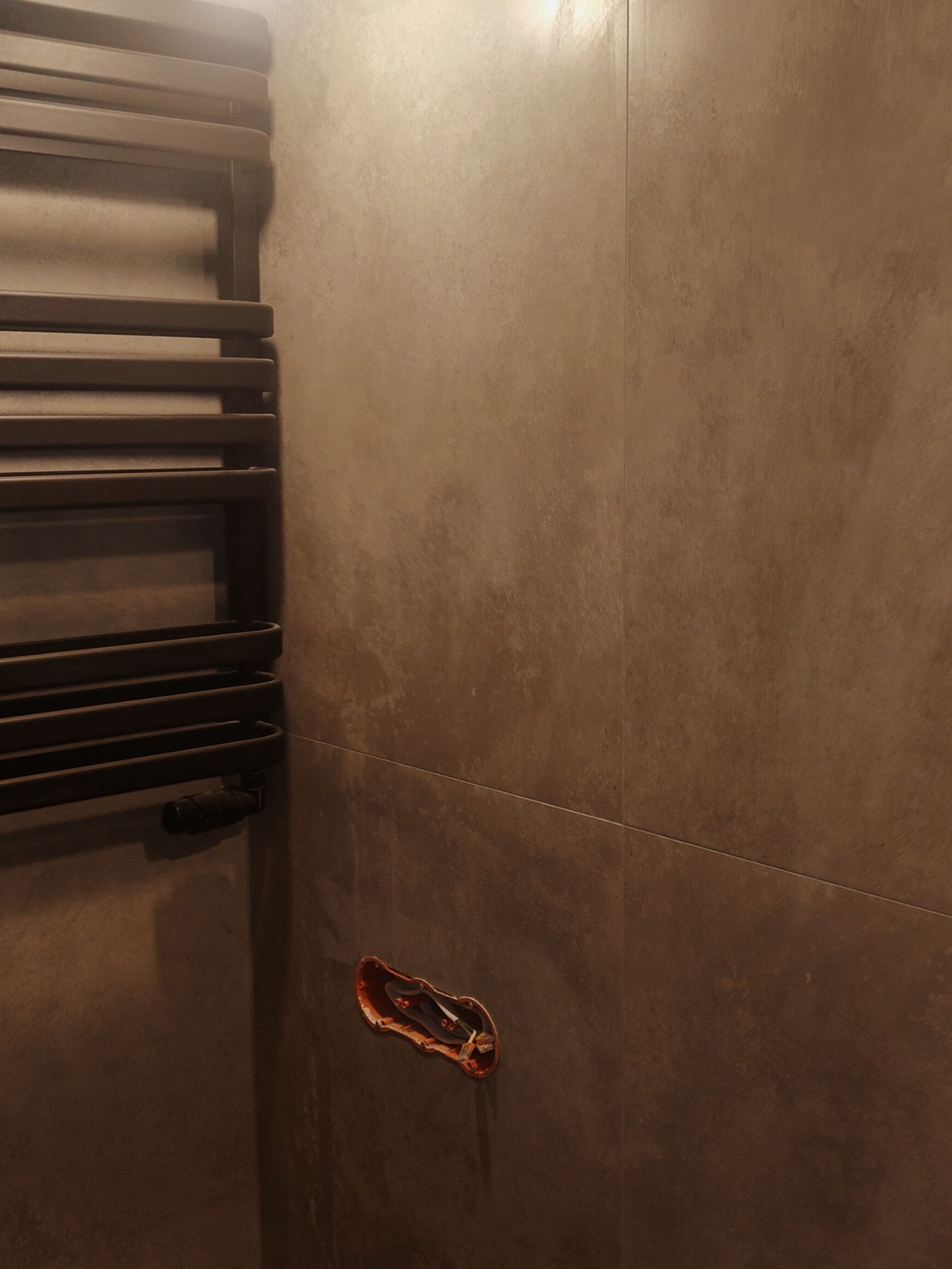 Brązowe płytki ścienne w łazience z zamontowanym grzejnikiem drabinkowym i niezabezpieczonym otworem w ścianie na instalację.