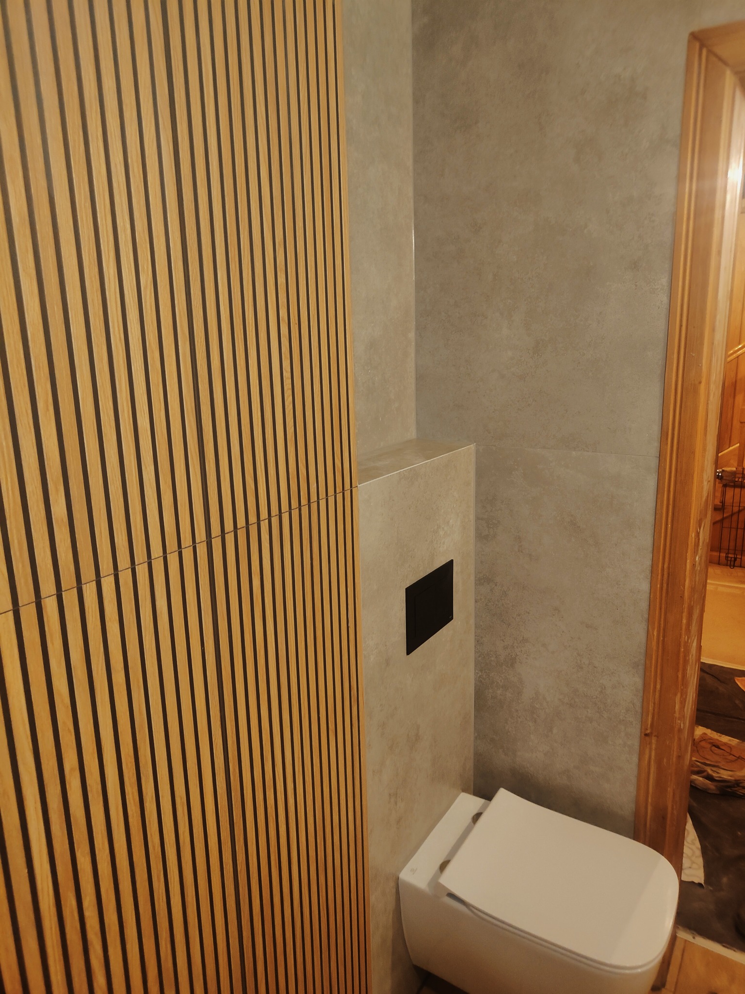 Nowoczesna łazienka z drewnianą ścianą z pionowych paneli, szare płytki imitujące beton i podwieszana toaleta z czarnym przyciskiem.