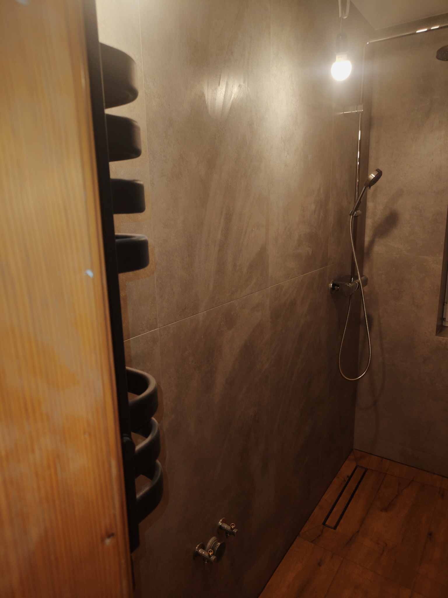 Nowoczesna kabina prysznicowa z drewnopodobną podłogą i szarymi płytkami imitującymi beton, widoczny grzejnik drabinkowy i minimalistyczna armatura.