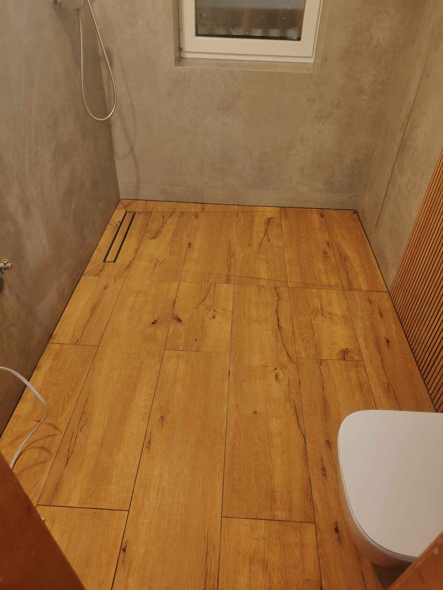 Wykończona łazienka z drewnopodobnymi płytkami na podłodze, szarymi płytkami na ścianach i odpływem liniowym w podłodze.