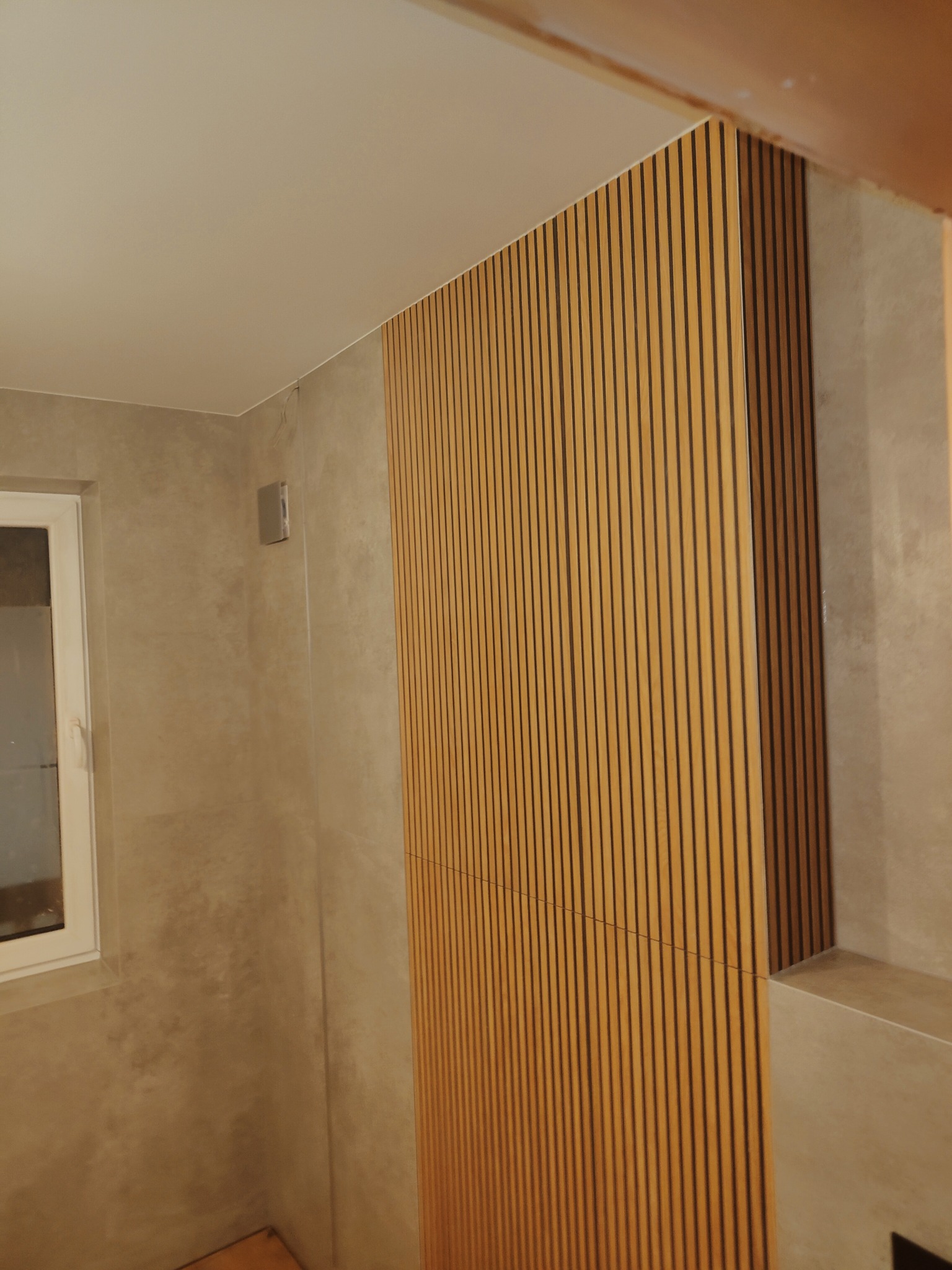 Ściana w łazience wykończona panelami z drewnianych listewek, sąsiadująca z płytkami imitującymi beton i białym oknem.