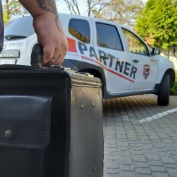 Partner Security Mariusz Kłosek - Mężczyzna z tatuażem na ramieniu trzyma czarną skórzaną aktówkę na tle białego samochodu dostawczego z napisem 'Partner' i logo firmy ochroniarskiej.