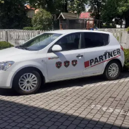 Biały samochód osobowy firmy ochroniarskiej Partner zaparkowany na brukowanym parkingu, z widocznymi logotypami firmy na karoserii i oznaczeniami na boku pojazdu.