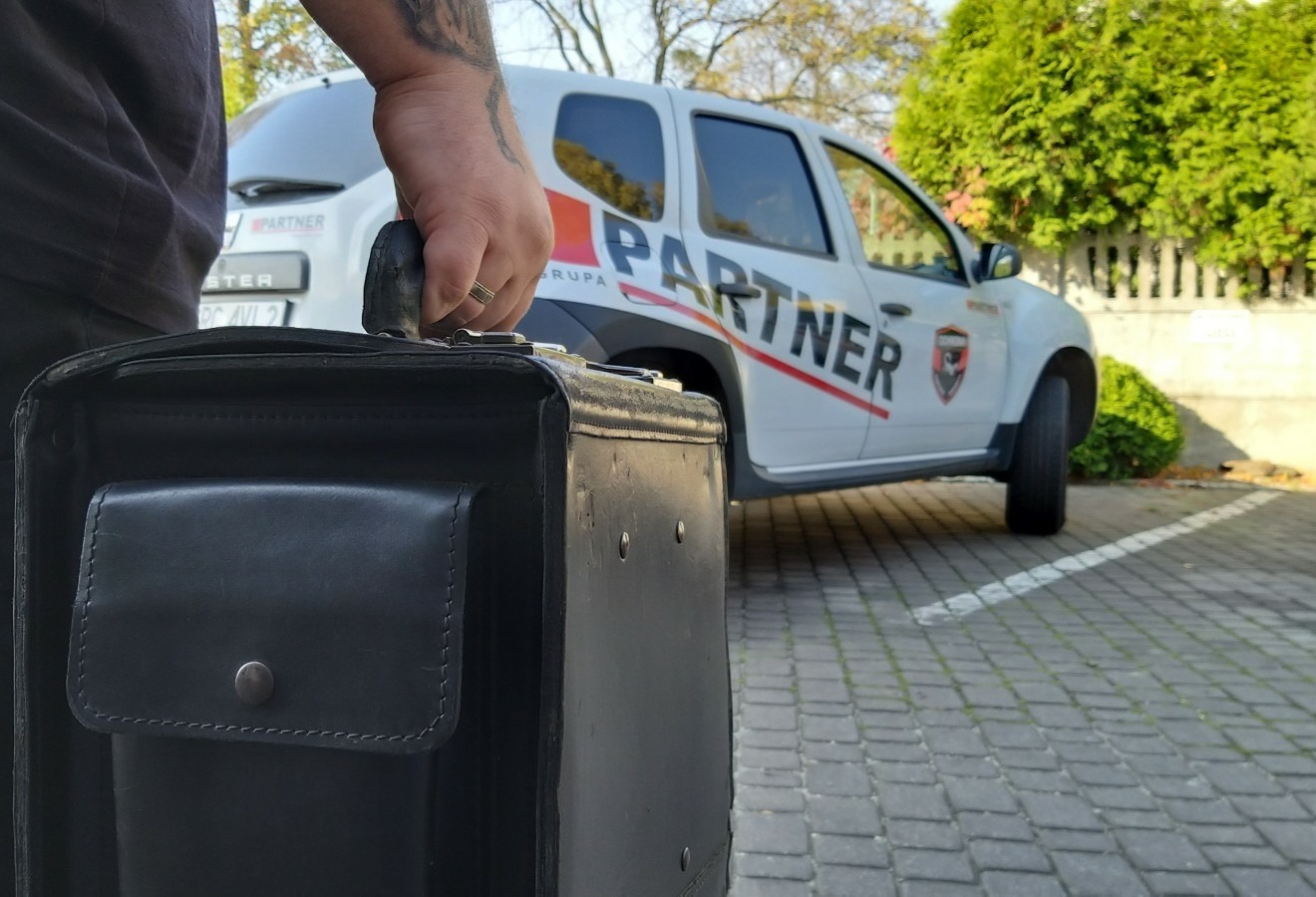 Mężczyzna z tatuażem na ramieniu trzyma czarną skórzaną aktówkę na tle białego samochodu dostawczego z napisem 'Partner' i logo firmy ochroniarskiej.