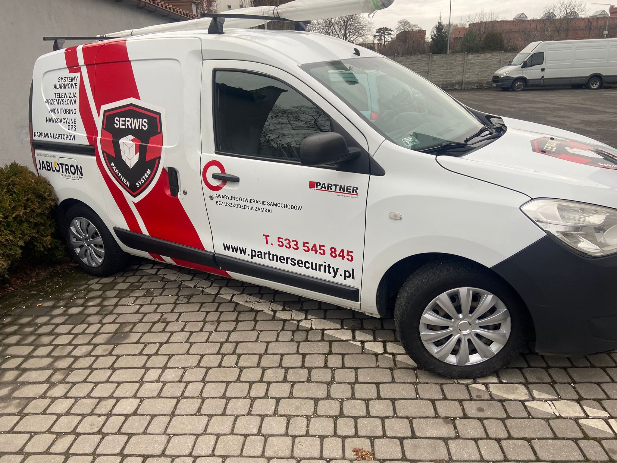 Biały samochód dostawczy z czerwonymi elementami graficznymi i logo firmy ochroniarskiej Partner System, zaparkowany na brukowanym parkingu. Widoczne numery telefonów i adres strony internetowej.