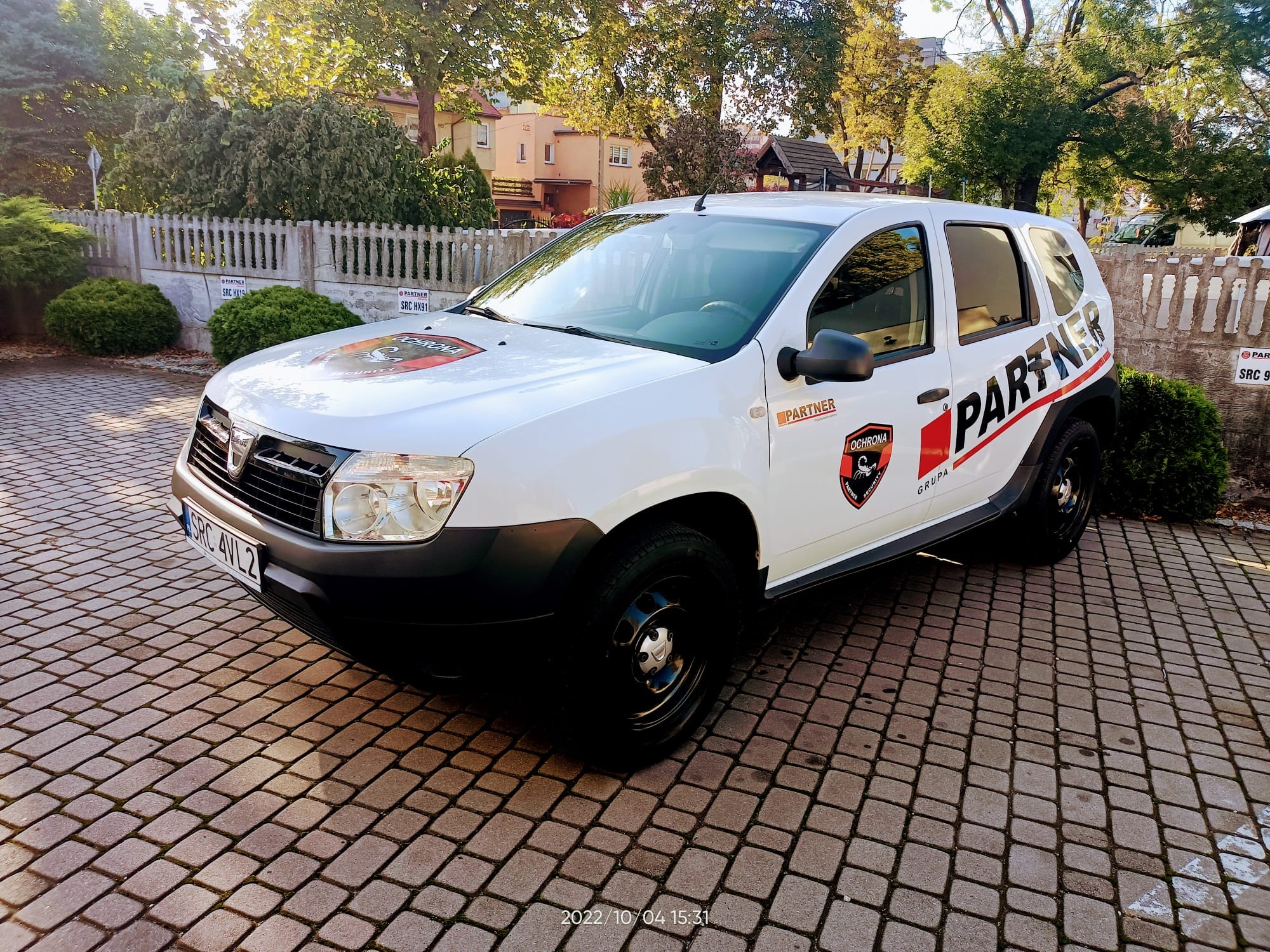 Biały samochód firmy ochroniarskiej Partner z logo firmy, zaparkowany na brukowanym podjeździe przed budynkiem i ogrodzeniem.