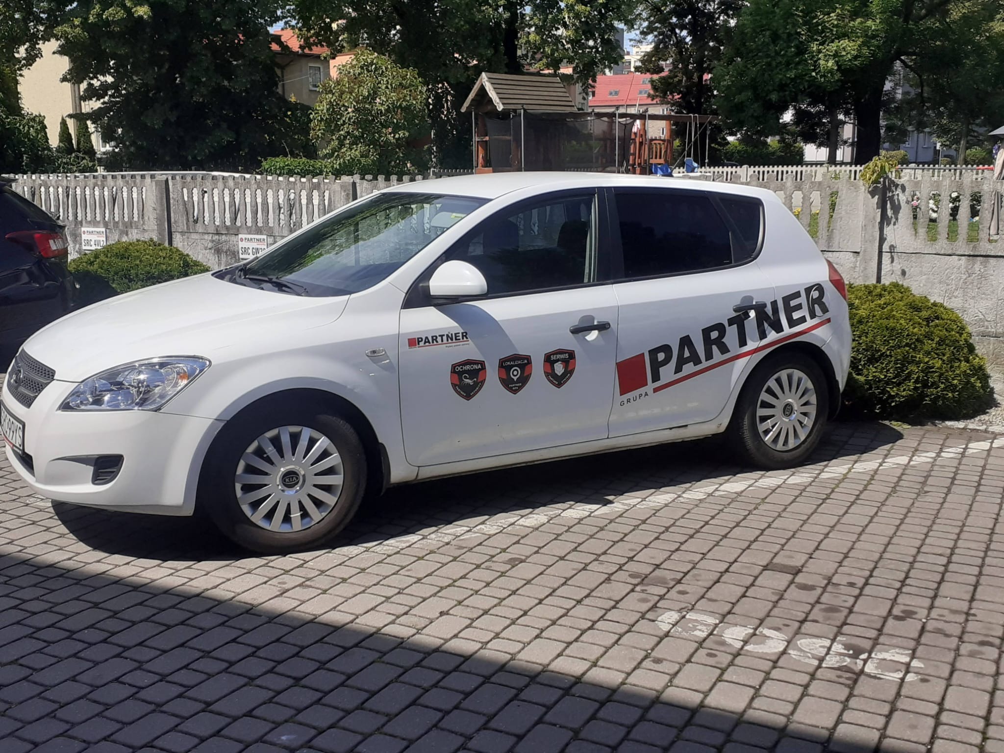 Biały samochód osobowy firmy ochroniarskiej Partner zaparkowany na brukowanym parkingu, z widocznymi logotypami firmy na karoserii i oznaczeniami na boku pojazdu.