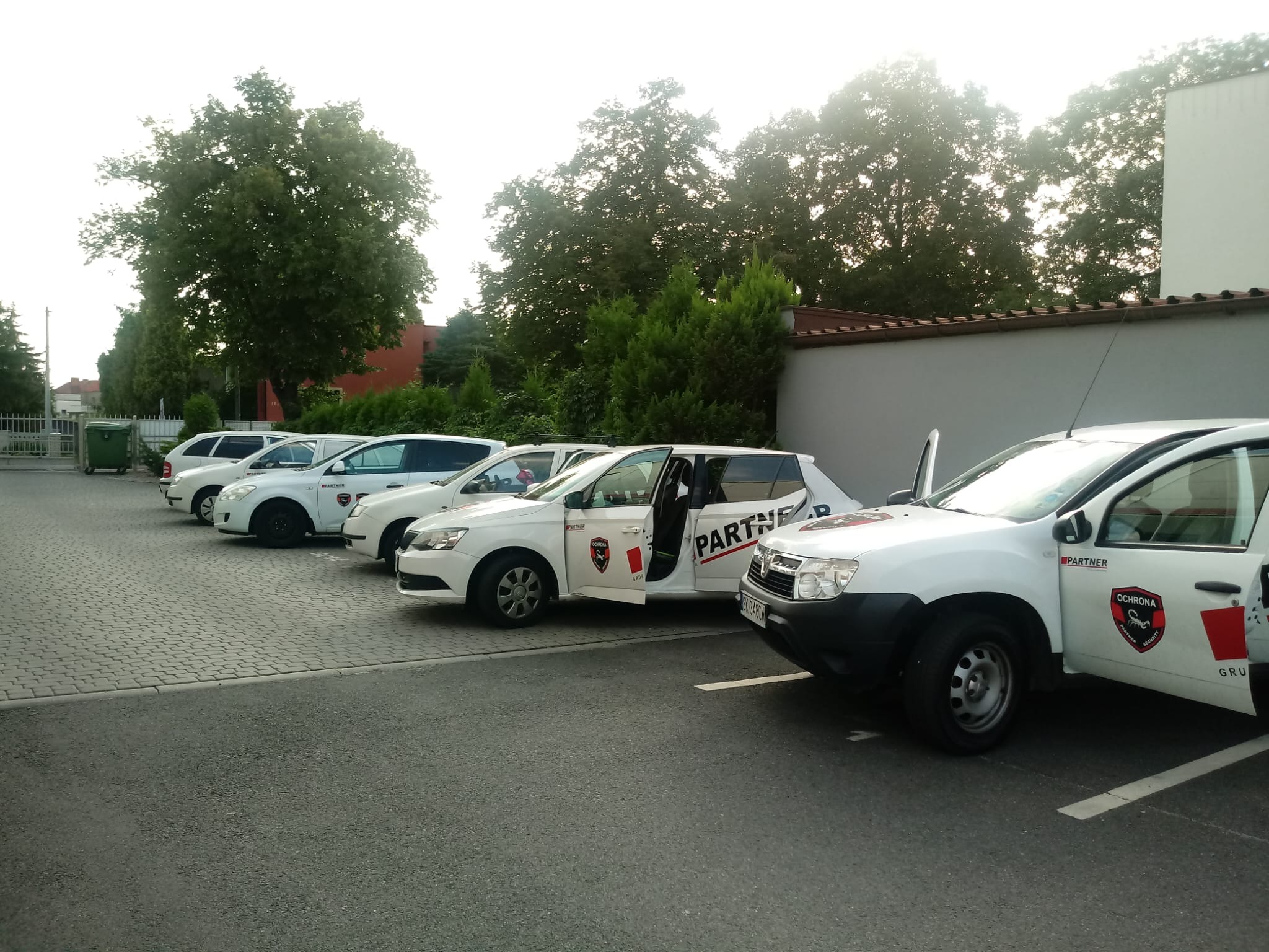 Szereg białych samochodów firmowych z logo 'Partner Ochrona' zaparkowanych na kostce brukowej, jeden z otwartymi drzwiami, na tle zieleni i fragmentu budynku.