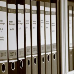 Rząd segregatorów Leitz z dokumentami księgowymi, oznaczonych rokiem 2015, uporządkowanych alfabetycznie według zakresu nazwisk klientów i dostawców, ustawionych na półce.