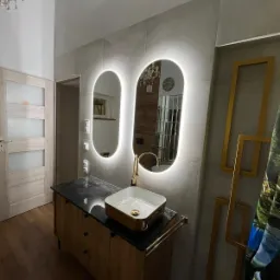 Nowoczesna łazienka z podświetlanymi lustrami w kształcie owali, umywalką nablatową z elementami złota na szafce z czarnym blatem i drewnianymi frontami, oraz złotymi ramkami dekoracyjnymi na ścianie.