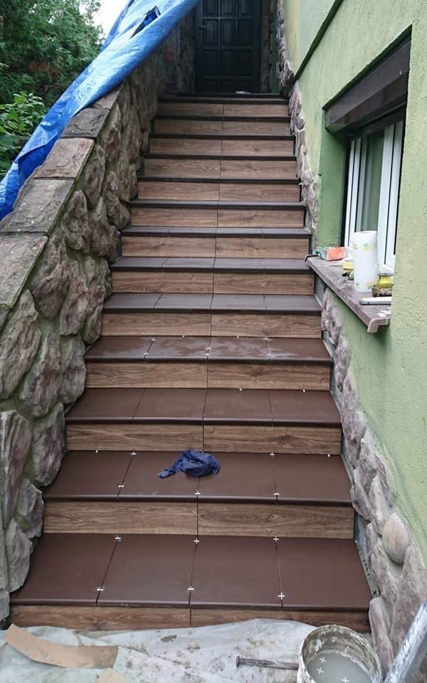 Wykończone schody zewnętrzne z brązowych płytek imitujących drewno, z widocznymi krzyżykami dystansowymi, z kamiennym wykończeniem po bokach i fragmentem okna w tle.