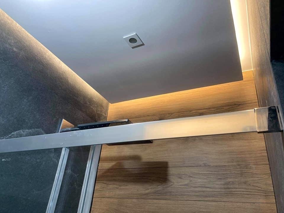 Szczegółowe ujęcie kabiny prysznicowej z szarymi płytkami imitującymi kamień i panelami imitującymi drewno, oświetlenie LED podsufitowe.