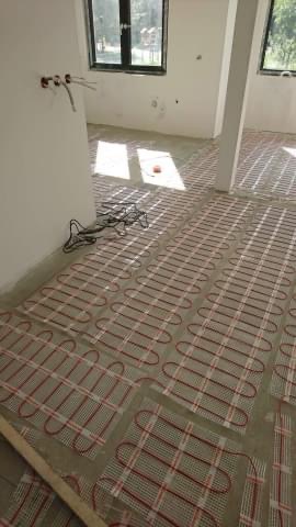 Rozłożona mata grzewcza na podłodze w surowym stanie, widoczne kable elektryczne i wystające ze ściany przewody, przygotowanie do wylewki.