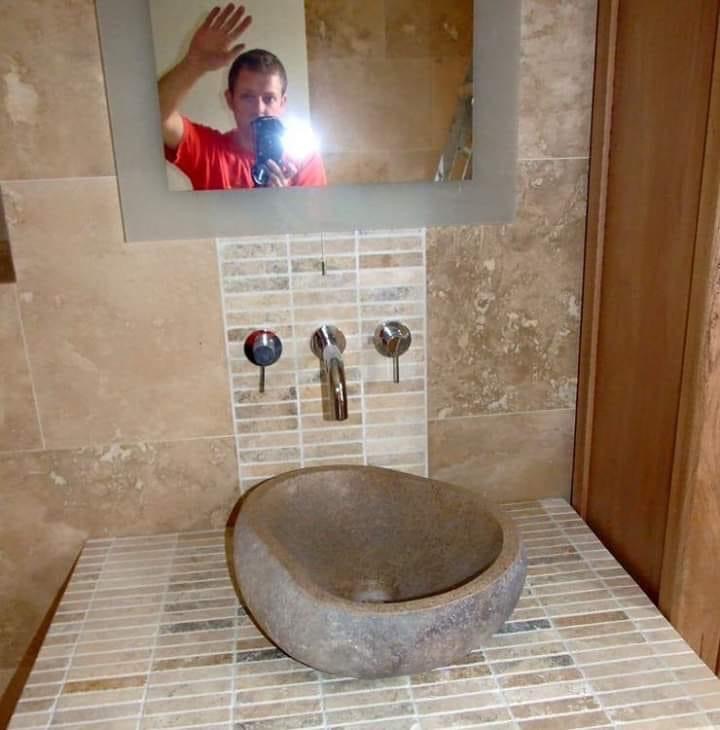 Umywalka z kamienia w łazience wykończonej mozaiką, widoczne trzy krany, odbicie fotografa w lustrze pozdrawiającego ręką.