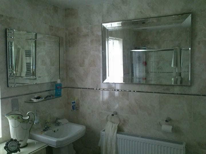 Wykończona łazienka z beżowymi płytkami, dwoma lustrami z fazowanymi krawędziami, umywalką na postumencie, grzejnikiem drabinkowym i dekoracyjnym wazonem.