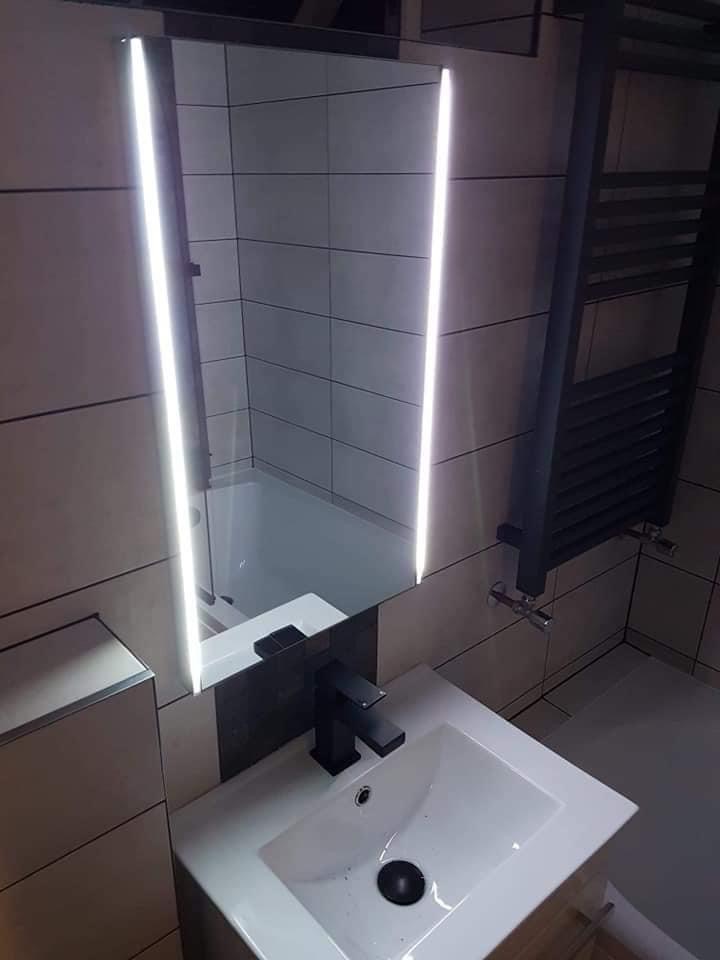 Nowoczesna łazienka z podświetlanym lustrem, czarną armaturą i grzejnikiem drabinkowym na tle płytek w odcieniach beżu i szarości.