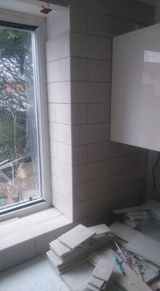 Ściana i parapet okna wyłożone jasnymi, prostokątnymi płytkami ceramicznymi w trakcie prac. Na podłodze stos płytek i narzędzia glazurnicze.