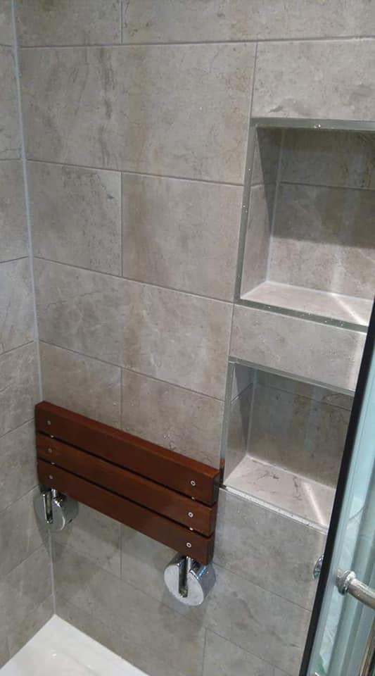 Zamontowane składane siedzisko prysznicowe z drewna w nowoczesnej łazience wyłożonej szarymi płytkami, z wnękami na kosmetyki obramowanymi szkłem.