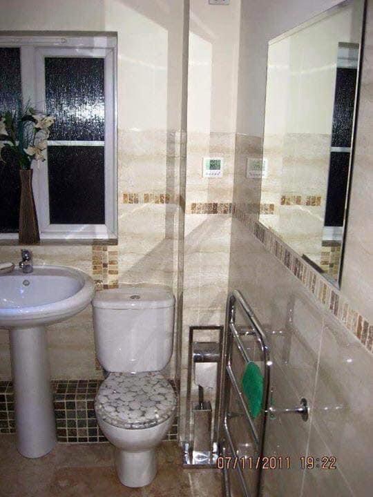 Wykończona łazienka w beżowych płytkach z dekoracyjnym pasem, umywalka na postumencie, toaleta z deską w mozaikę, grzejnik drabinkowy z zielonym ręcznikiem, lustro w ramie.