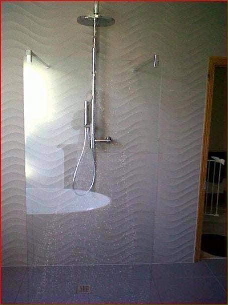 Nowoczesna kabina prysznicowa z deszczownicą i szklaną ścianką, wykończona płytkami z falistym wzorem w odcieniach szarości, z odpływem liniowym w podłodze.