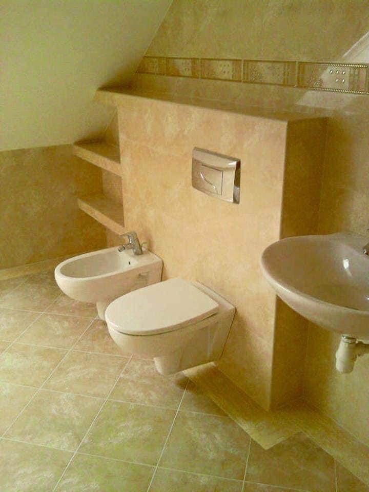 Wykończona łazienka na poddaszu z beżowymi płytkami, wiszącą toaletą, bidetem i umywalką, zabudowana ścianka z półkami.