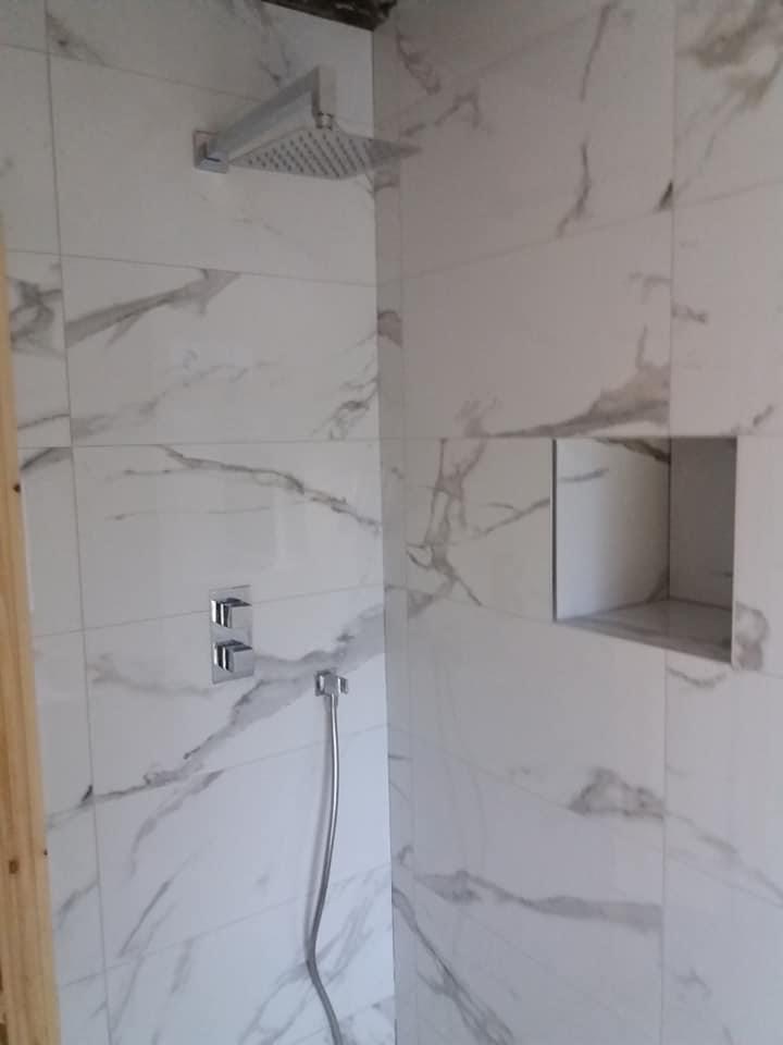 Wykończona kabina prysznicowa z marmurowymi płytkami, deszczownicą i wnęką na kosmetyki, widoczny fragment drewnianej ramy.