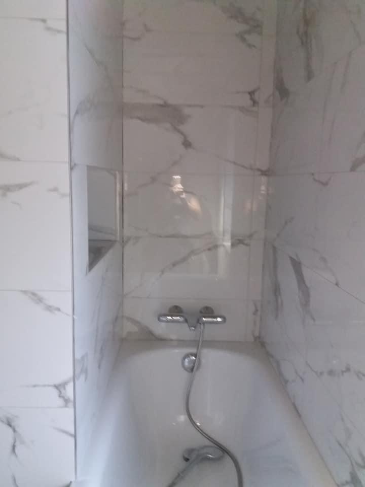 Biała wanna obudowana marmurowymi płytkami z szarymi żyłami, bateria wannowa, wnęka w ścianie wykończona płytkami.