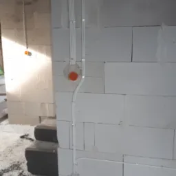 Surowe wnętrze w trakcie prac: białe ściany z bloków, pionowo poprowadzone kable elektryczne w białych rurkach mocowanych klamrami, pomarańczowe puszki instalacyjne w ścianie, widoczne ślady zaprawy.