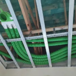 Zielone, karbowane rury wentylacyjne ułożone pod sufitem podwieszanym, widoczne elementy konstrukcji drewnianej i metalowej.