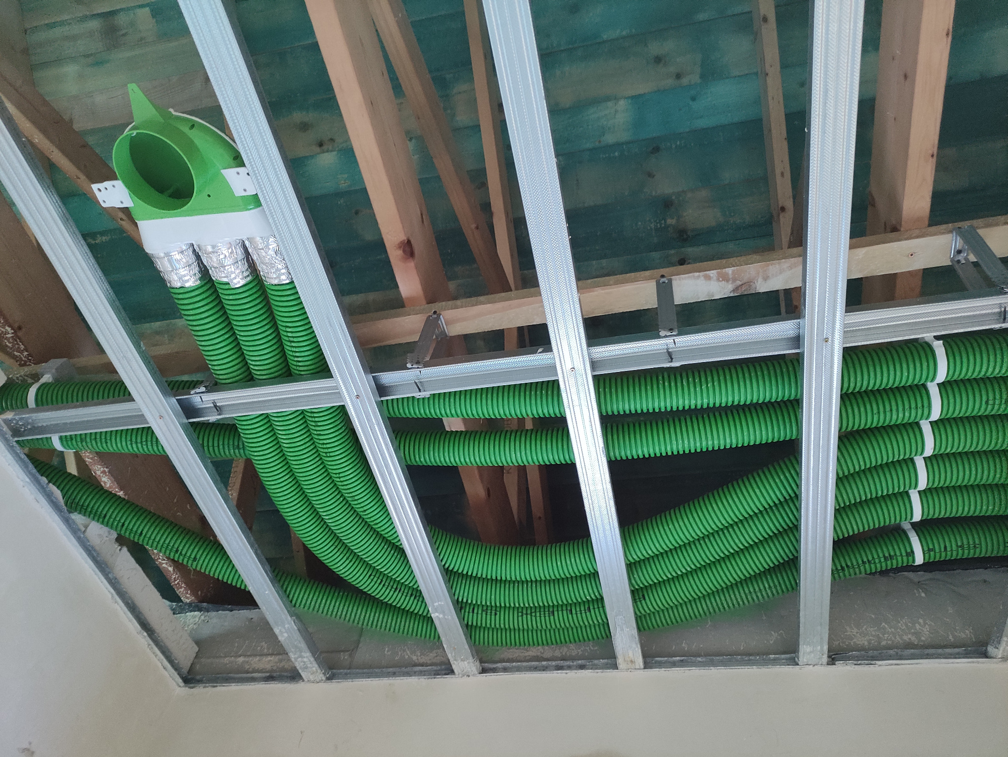 Zielone, karbowane rury wentylacyjne ułożone pod sufitem podwieszanym, widoczne elementy konstrukcji drewnianej i metalowej.