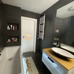 Nowoczesna łazienka w ciemnych kolorach z białą wanną, umywalką nablatową na drewnianym blacie i grzejnikiem drabinkowym; na ścianie wnęki z kosmetykami i dekoracyjny dywanik.