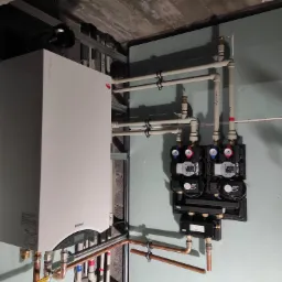 Nowoczesny piec gazowy Termet zamontowany w kotłowni, z widocznymi rurami miedzianymi i plastikowymi, oraz dwoma czarnymi rozdzielaczami z pompami obiegowymi i manometrami z czerwonymi...