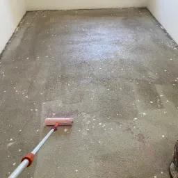 Wyrównywanie wylewki samopoziomującej w pomieszczeniu za pomocą wałka kolczastego, widoczne białe odpryski i ślady różowej farby.