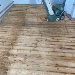 Euro instal parkiety - Zielona szlifierka do parkietu Hummel stoi na częściowo wyszlifowanej drewnianej podłodze; widoczne ślady pyłu i różnica w odcieniu drewna przed i po szlifowaniu.