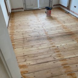 Euro instal parkiety - Drewniana podłoga po częściowym nałożeniu bejcy, widoczne ślady po pędzlu, puszka z bejcą i zabezpieczony grzejnik w tle.