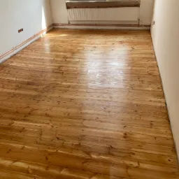 Świeżo polakierowana drewniana podłoga z widocznymi miedzianymi rurami grzewczymi przy ścianie i grzejnikiem w tle, w trakcie remontu pomieszczenia.